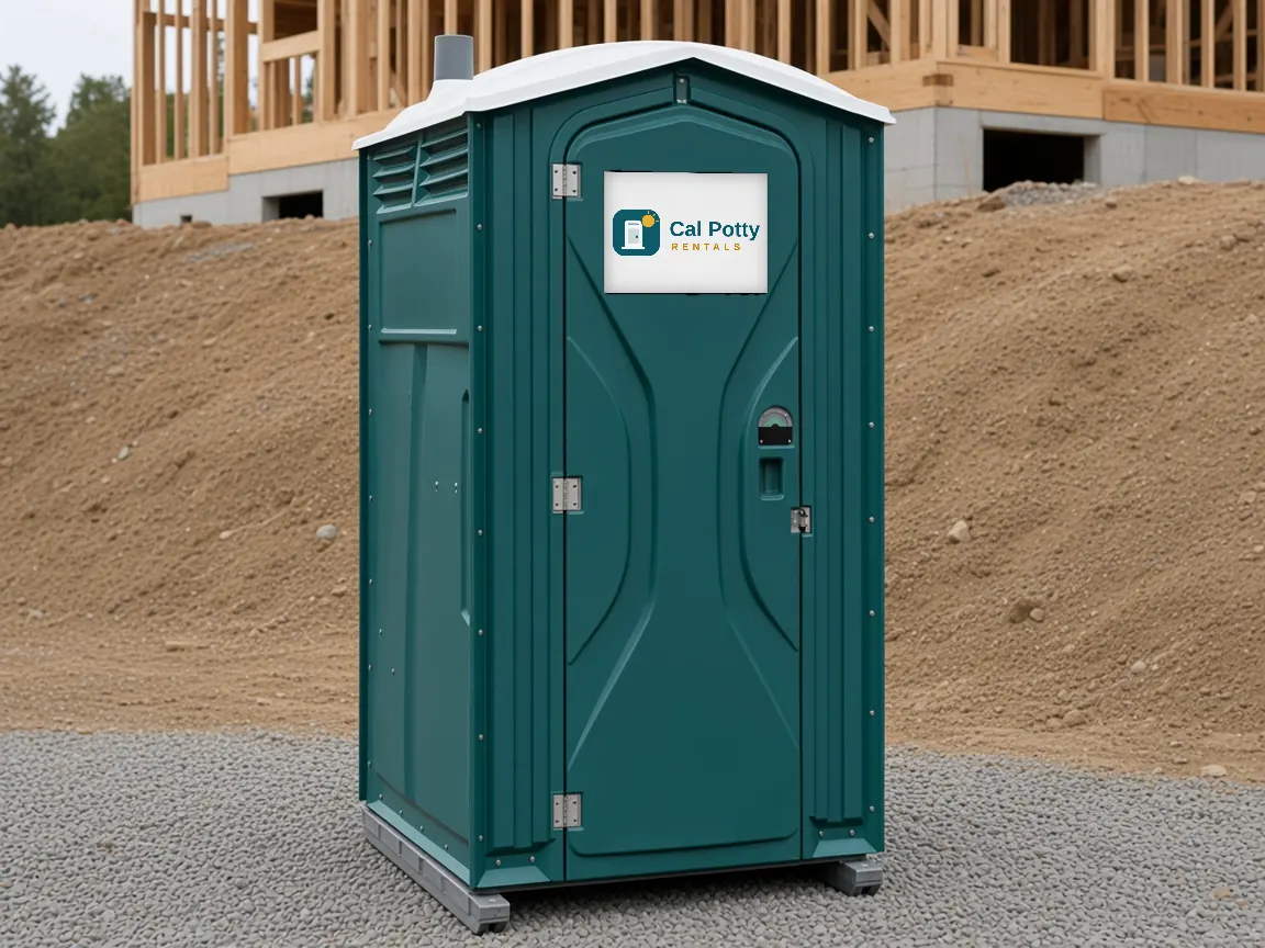 A teal porta potty on a gravel pad in front of a dirt mound and a wood-framed building